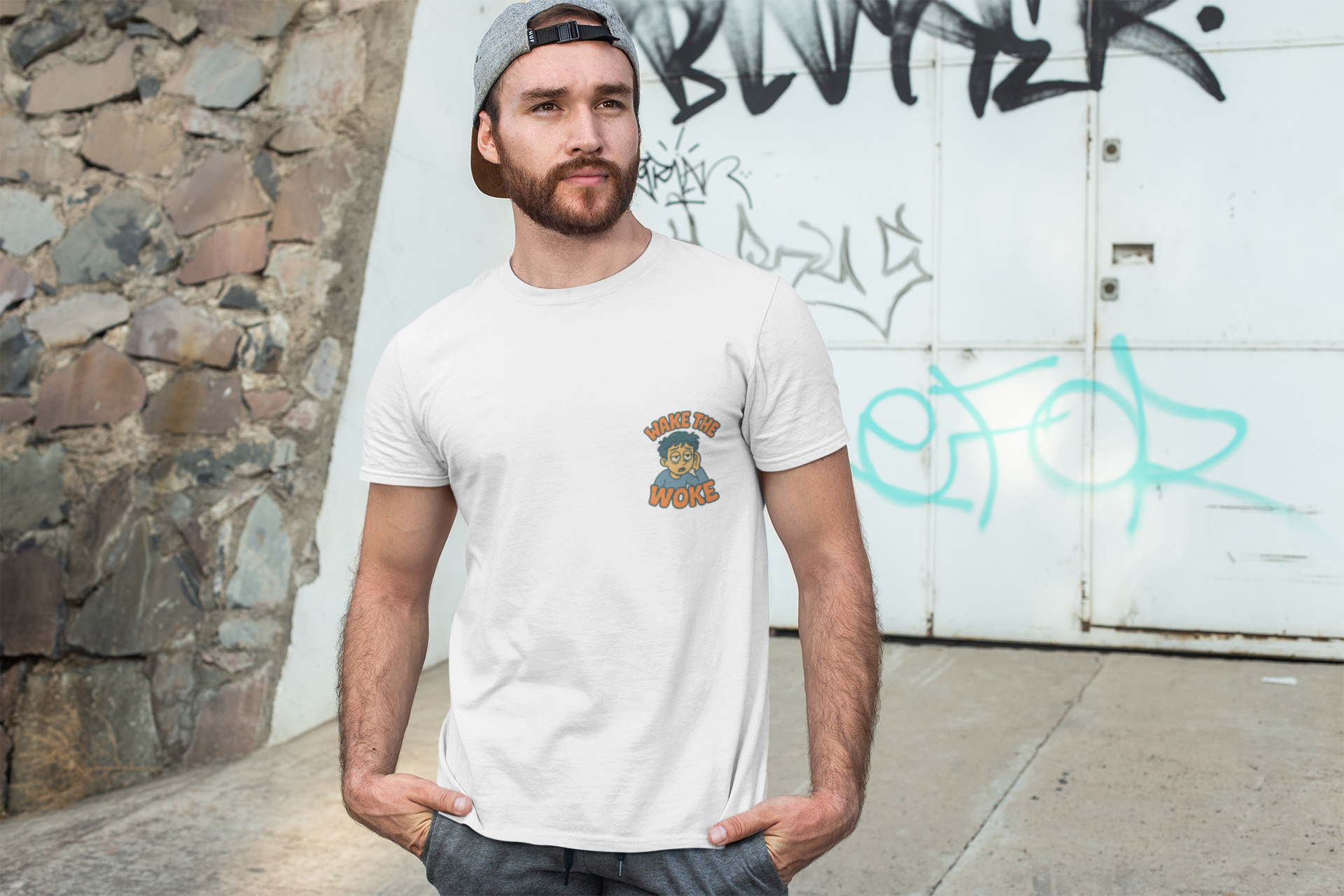 A man wearing a white crewneck t-shirt with a small graphic on the left chest, paired with a gray cap and jeans, standing outdoors against a graffiti-covered white wall.